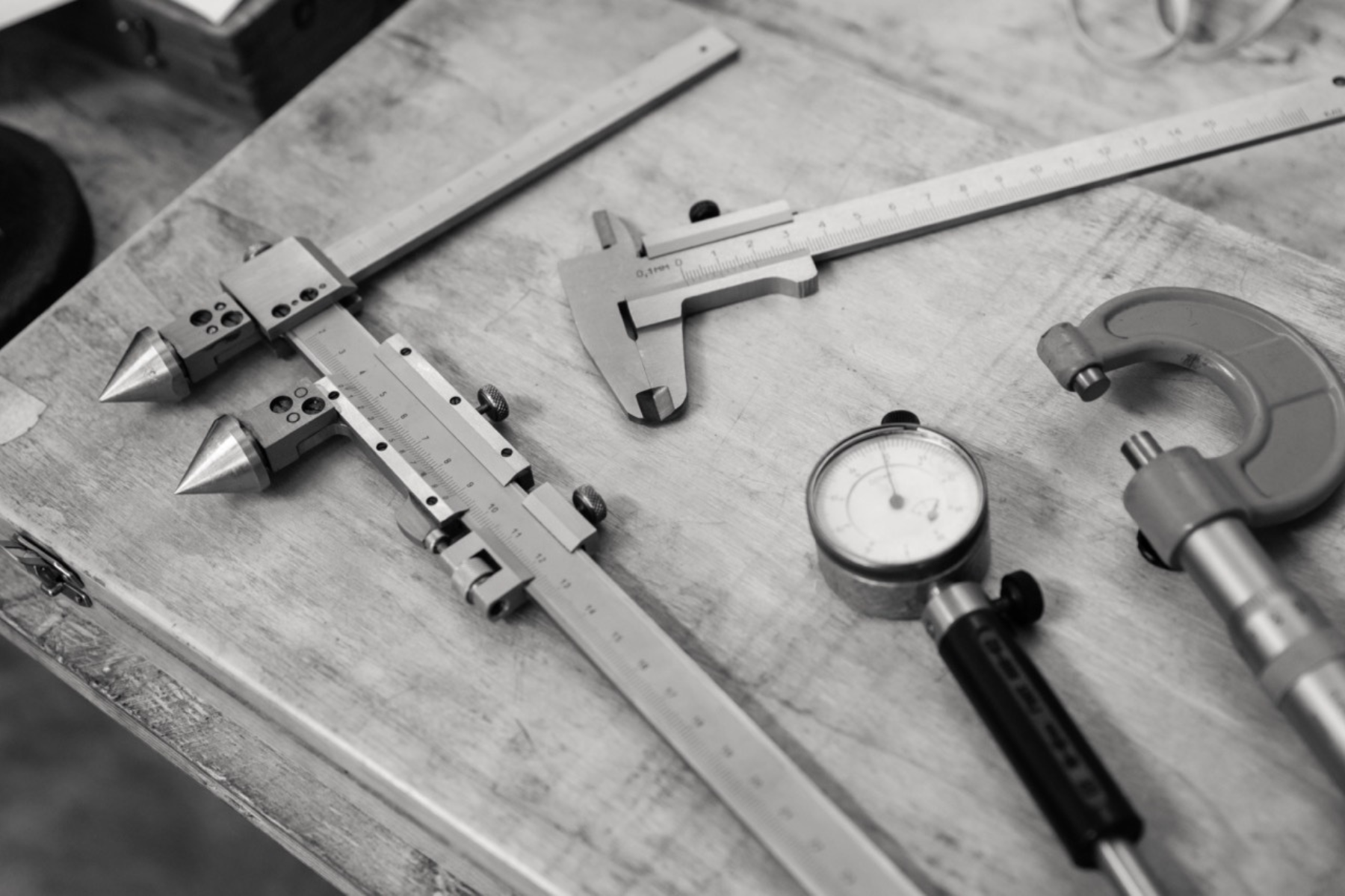 The image is a black-and-white photograph of a workbench displaying various precision measuring tools. It includes two calipers with pointed tips, a Vernier caliper with a detailed scale, a dial indicator with a circular gauge, and a micrometer with a curved frame. The tools are arranged on a worn wooden surface, suggesting a workshop or machining environment.
