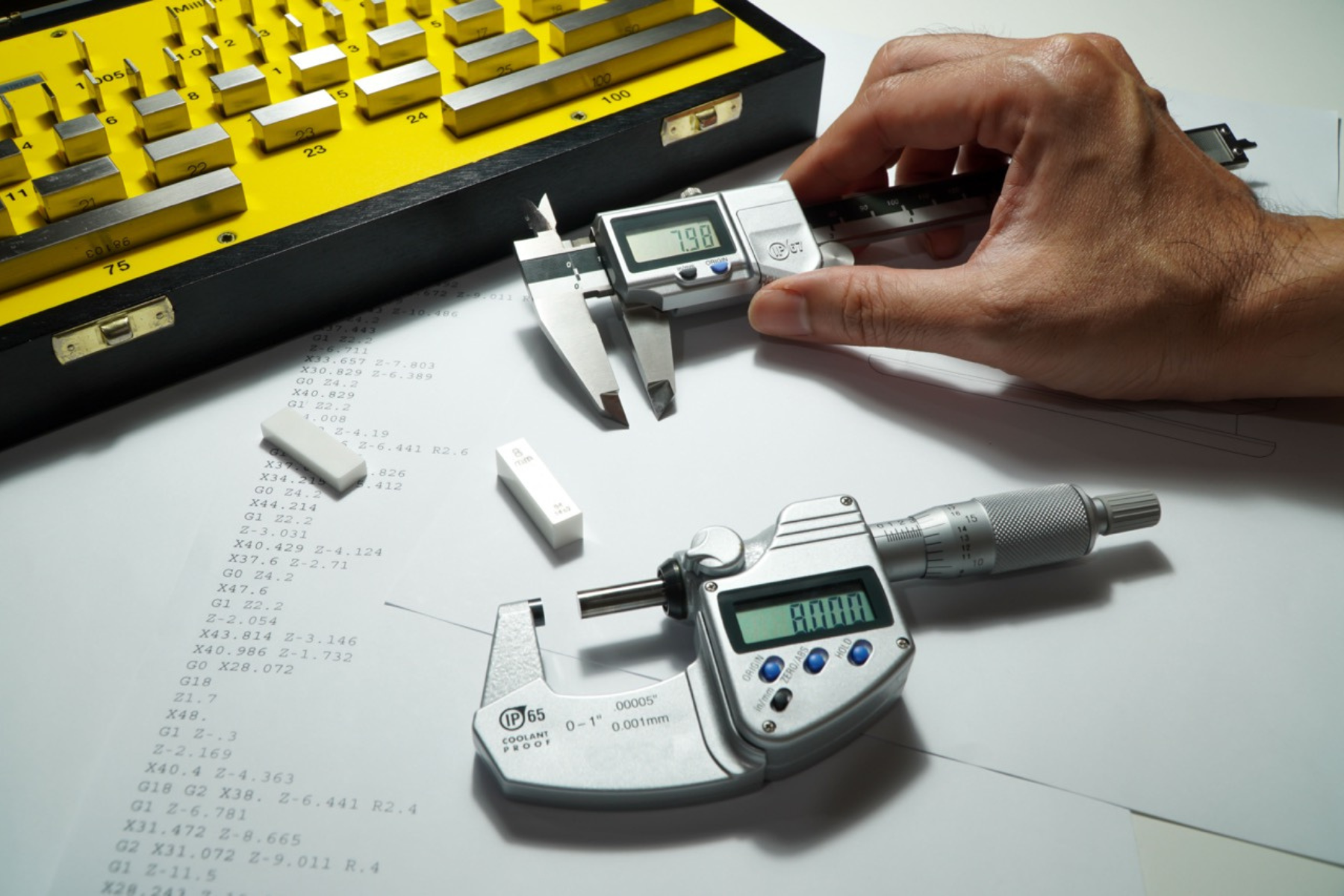 Hands measure metal blocks with a micrometer and digital caliper, with calibration standards nearby.