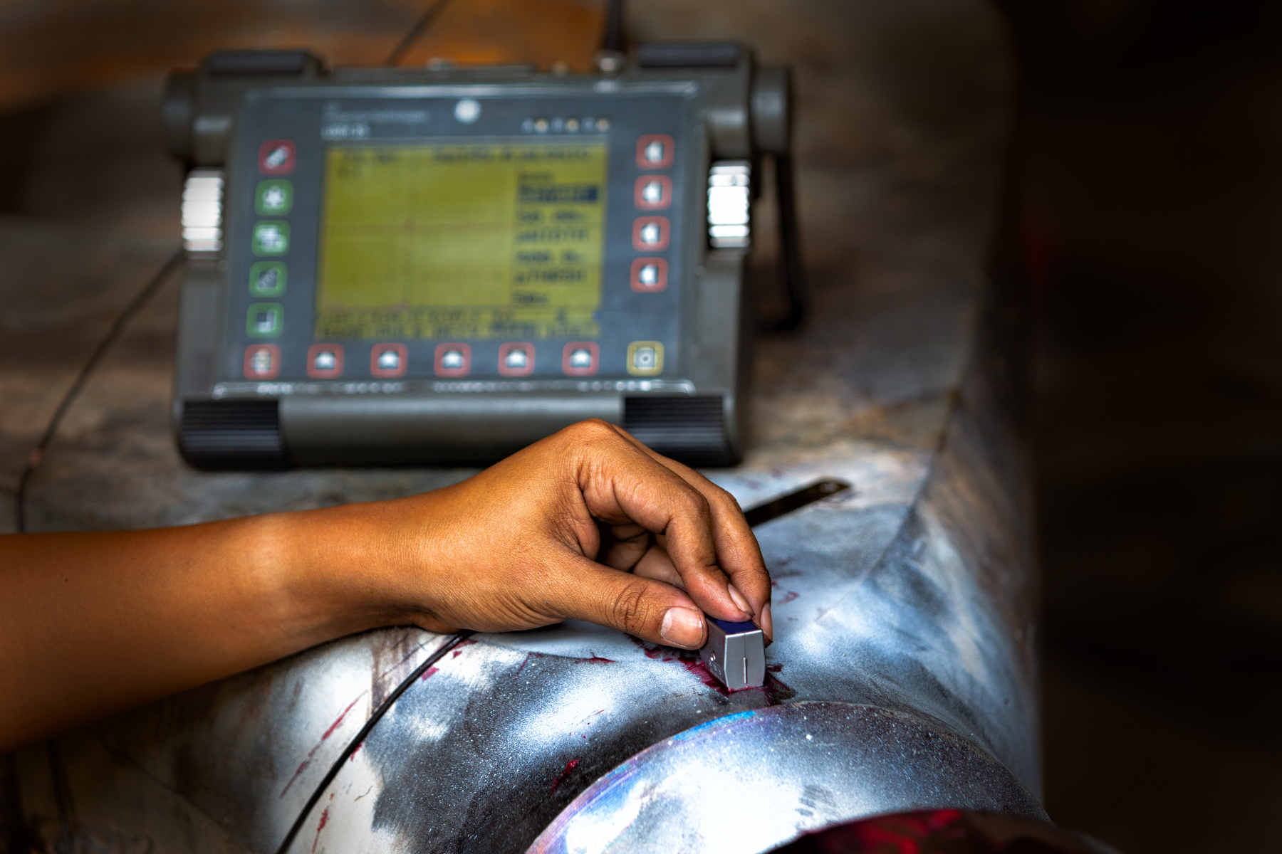 A close-up of a hand using a device to measure a metal pipe, with a portable screen displaying data in an industrial setting.