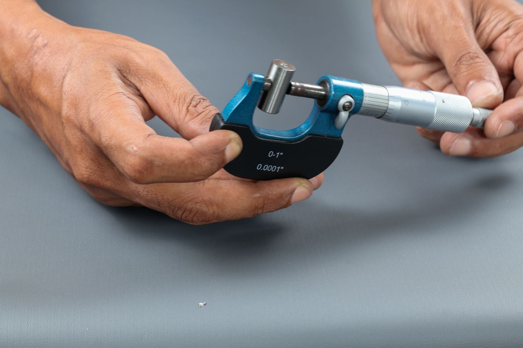 Hands holding a blue and silver micrometer, adjusting it to measure a small cylindrical metal object, against a gray work surface.