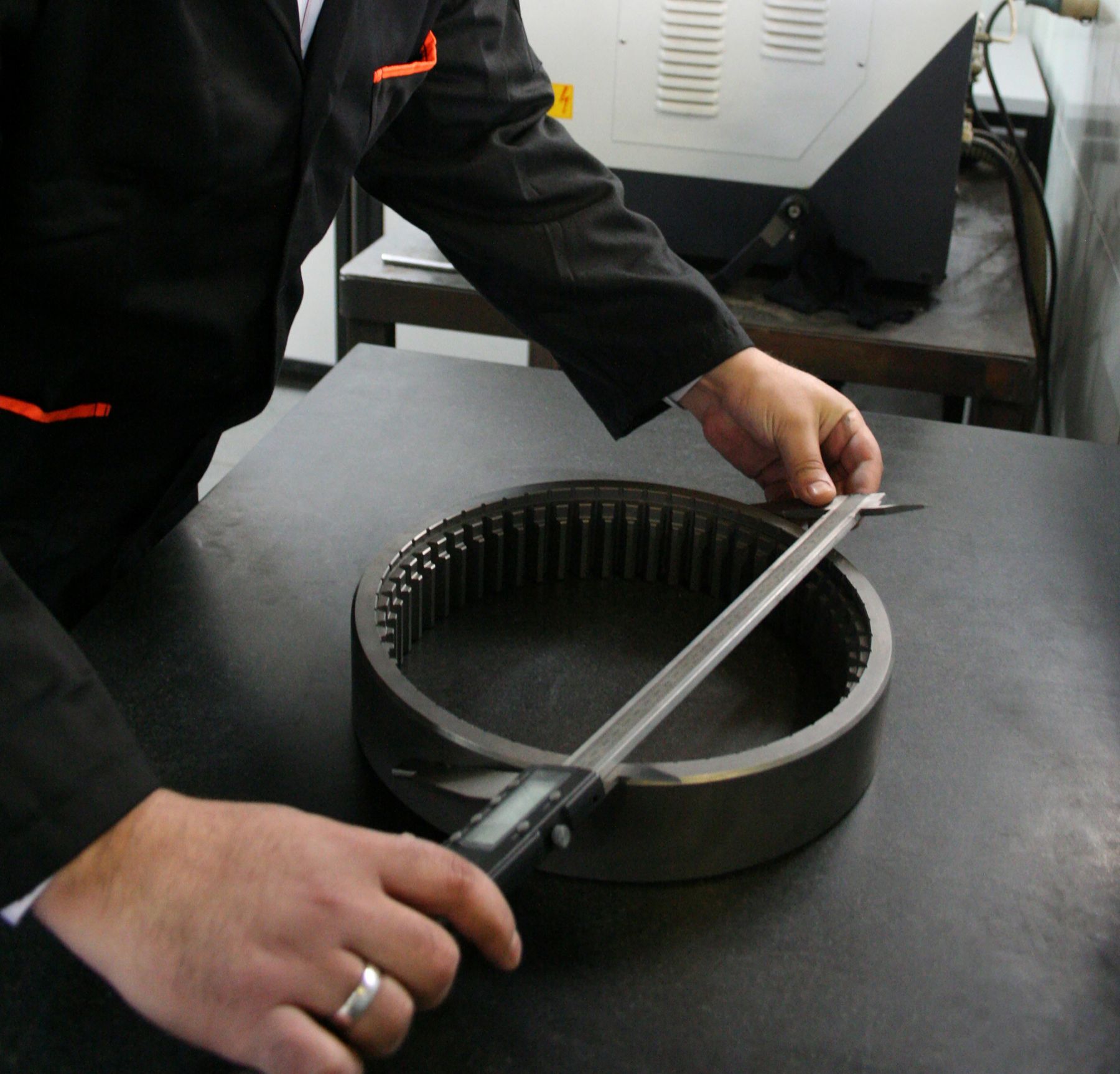A person using a digital caliper to measure the inner diameter of a large circular metal component with teeth along its inner edge, placed on a dark surface in a workshop.