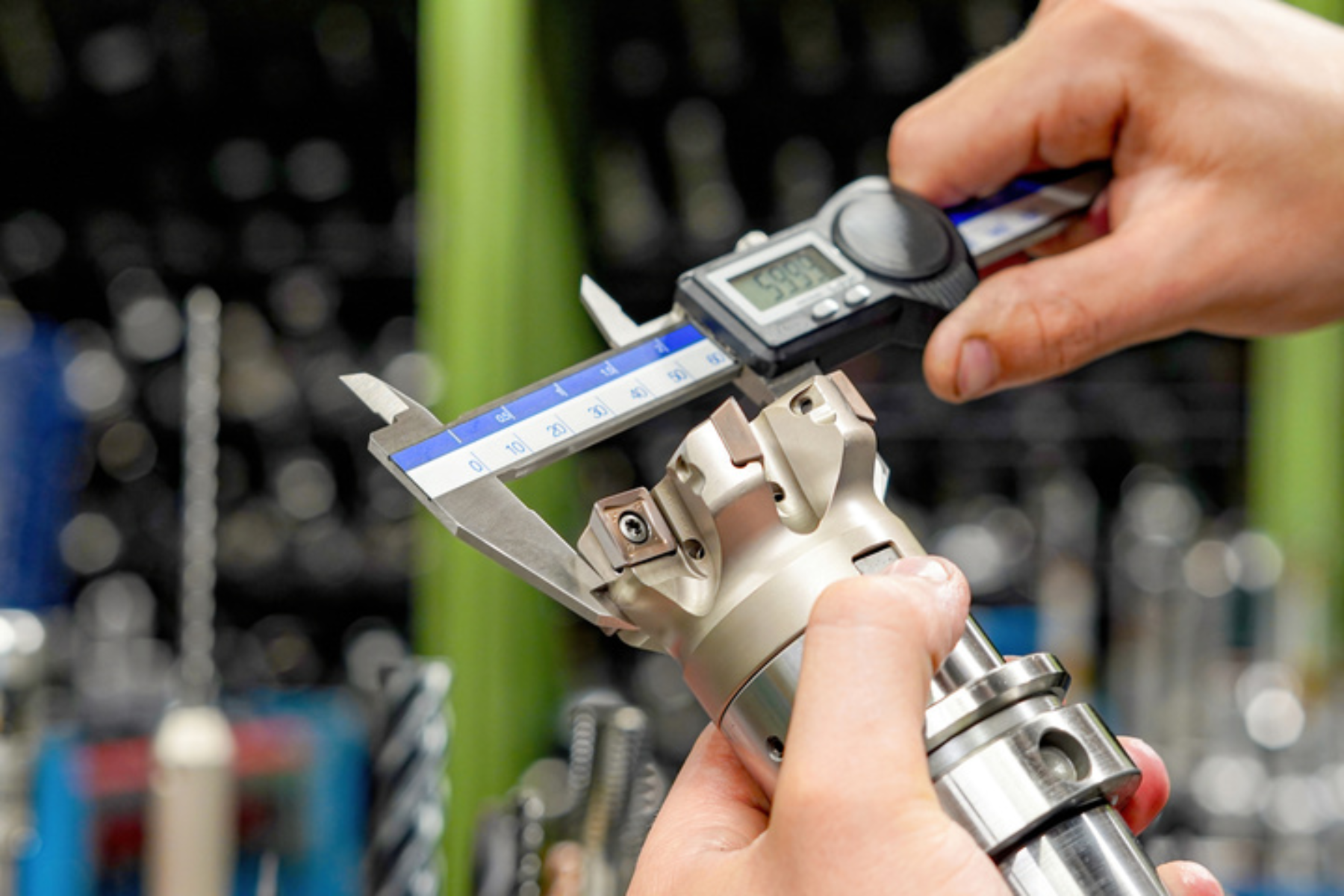 Hands use a digital caliper to measure a complex metal tool component, showing precision inspection work in manufacturing.