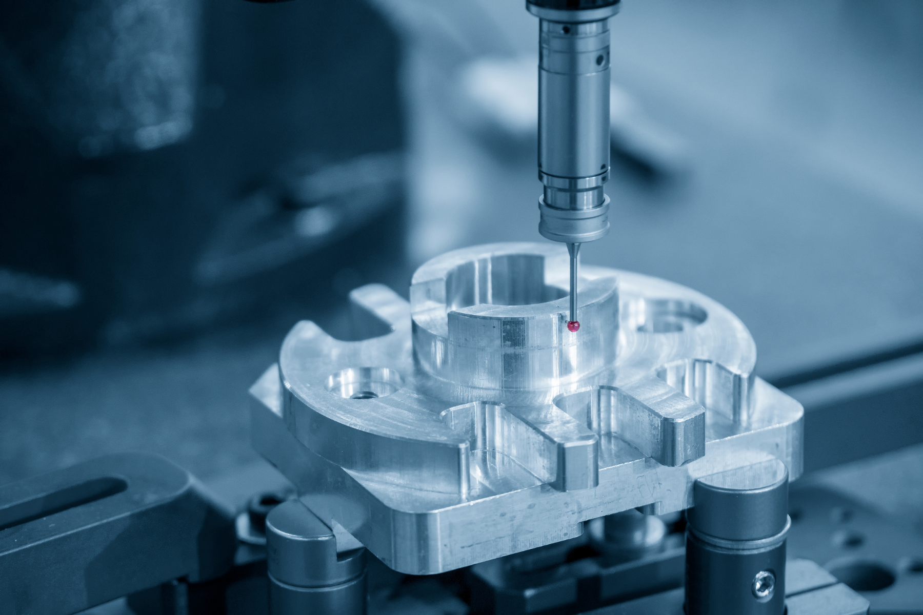 Blue-toned close-up of a measuring probe touching or hovering just above a shiny machined metal part mounted in a fixture. The part has circular and radial features, and the image highlights precision dimensional inspection.