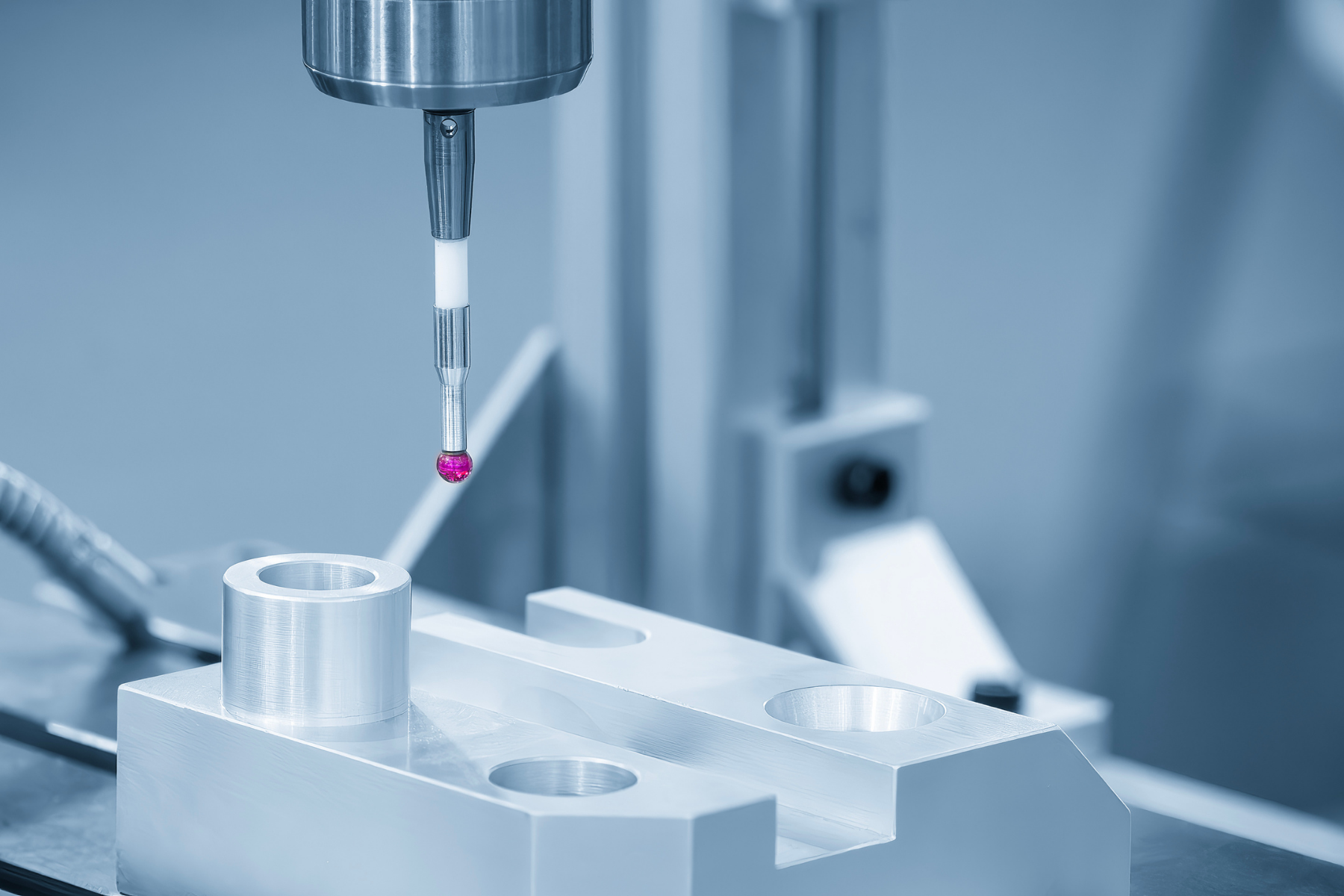 A close-up industrial image of a coordinate measuring machine probe with a small pink ruby tip positioned above a machined metal component. The scene has a cool blue tone and emphasizes precision inspection in a metrology or manufacturing setting.